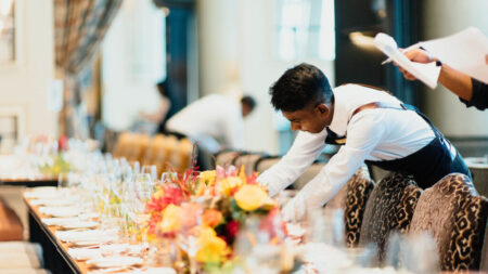 A waiter is leaning over a long table which has plates and flowers on it.