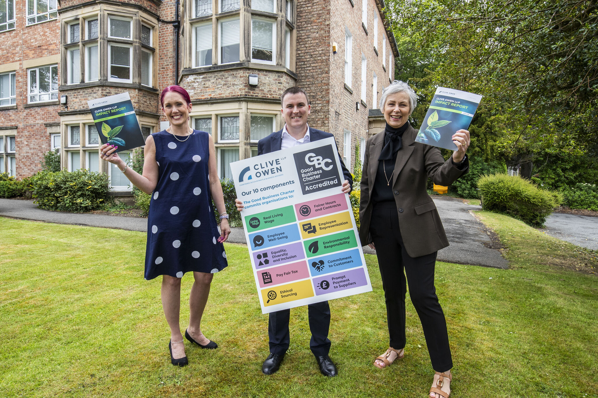 Three people stand outside a building, holding a sign displaying the components of the Good Business Charter and impact report pamphlets.