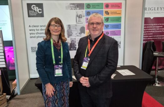 Two professionals stand in front of a booth displaying the Good Business Charter, promoting responsible business practices.