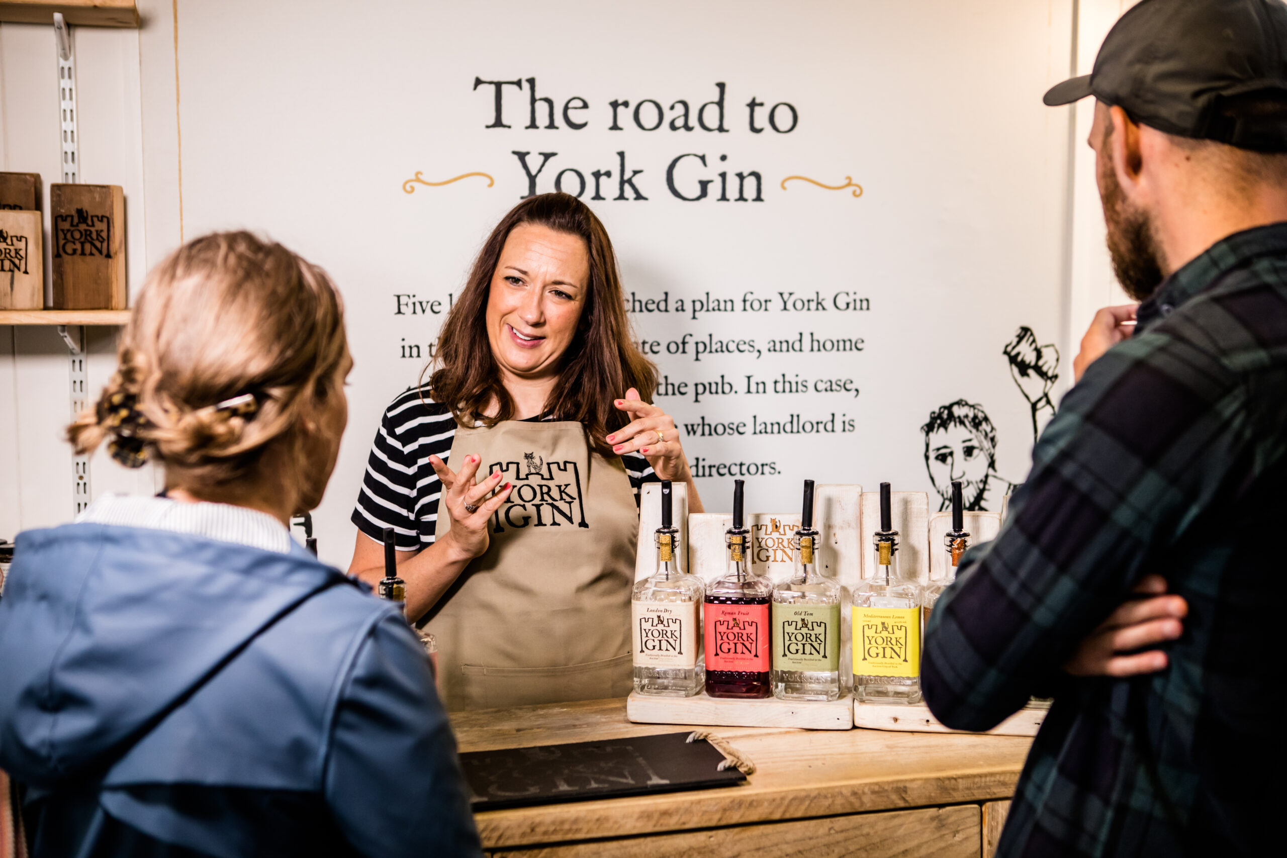 A bartender at a York Gin booth explains products while two customers listen attentively, surrounded by bottles of various gin flavours.