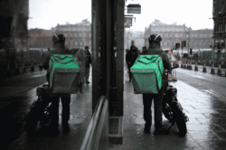 Food delivery rider with a green backpack and motorcycle on a wet, overcast day.