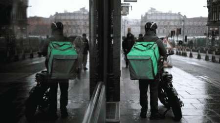 Food delivery rider with a green backpack and motorcycle on a wet, overcast day.